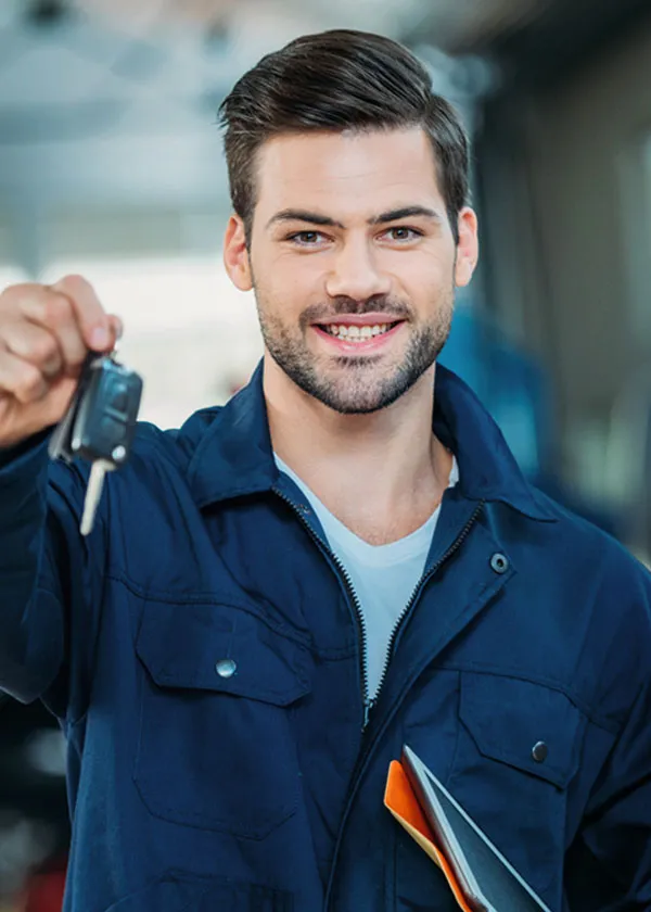 Smiling mechanic in a blue uniform holds out car keys and a clipboard inside an auto repair shop.